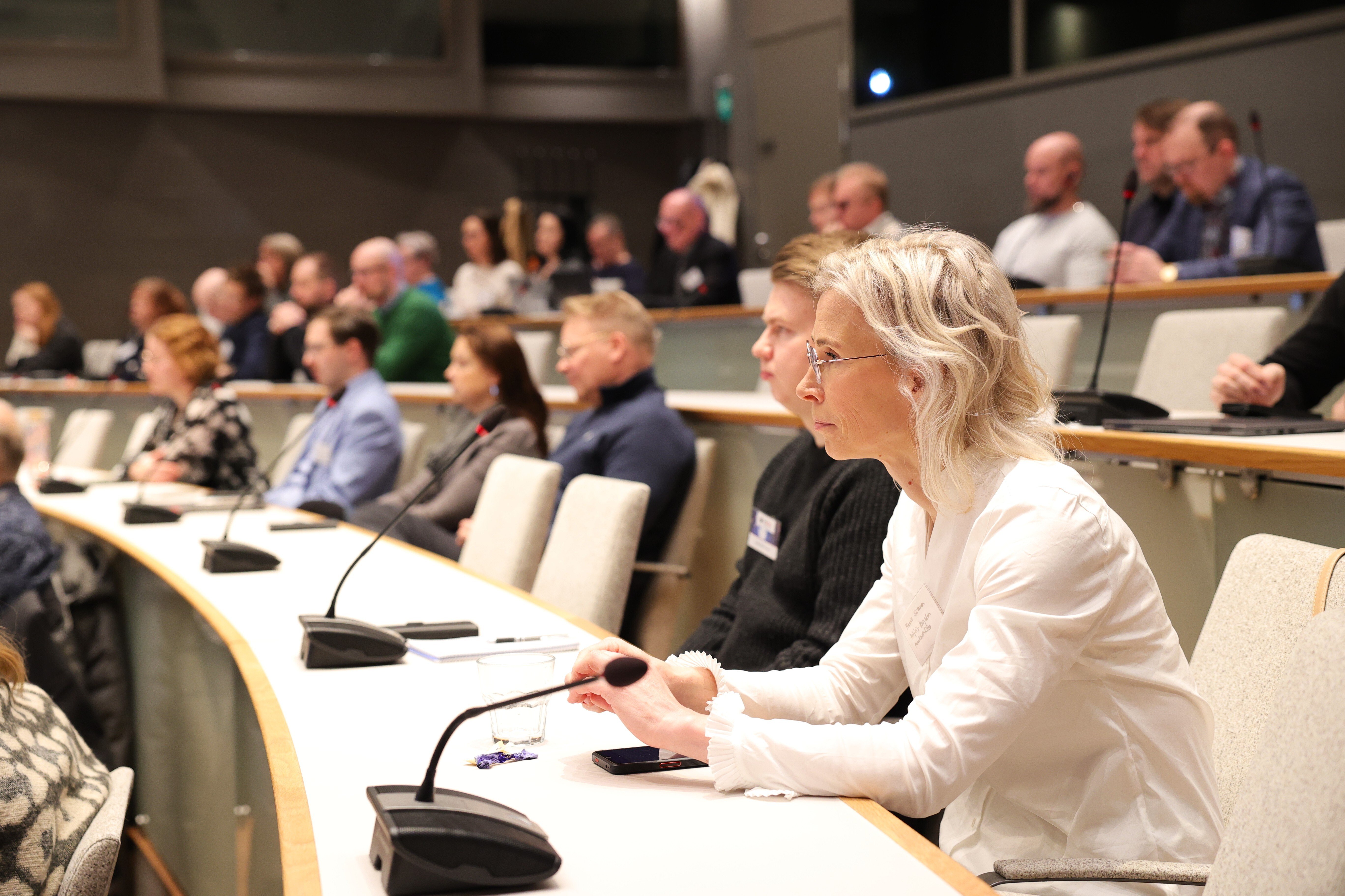 Joukko ihmisiä istuu auditoriossa ja katsoo eteensä keskittyneenä.