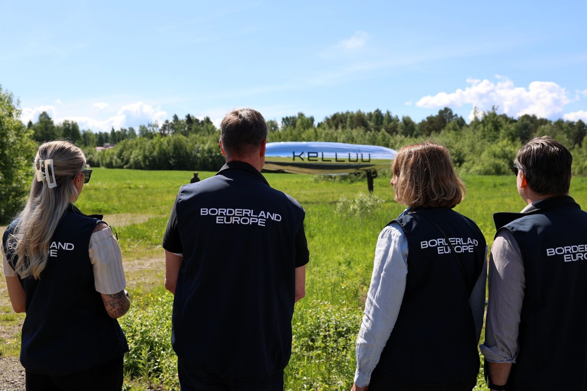 Neljä henkilöä selin kameraan päin ja katsovat hopeista, pitkulaista ilmalaivaa.
